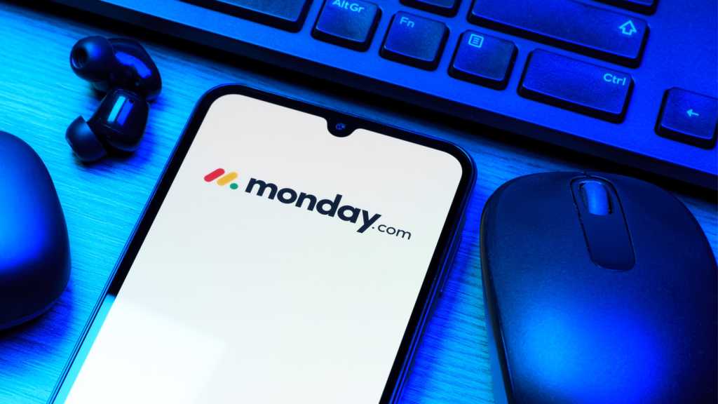 A photograph of a smartphone showing the Monday.com logo on a desk with blue lighting surrounded by a keyboard, mouse and wireless earphones.